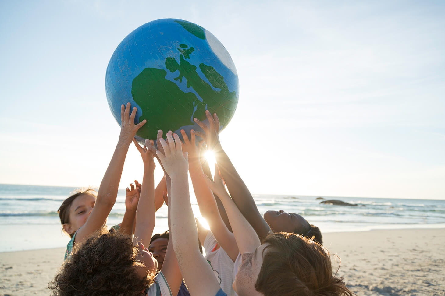 5 Kinder halten gemeinsam am Sandstrand eine Weltkugel, durch ihre Hände scheint die Sonne