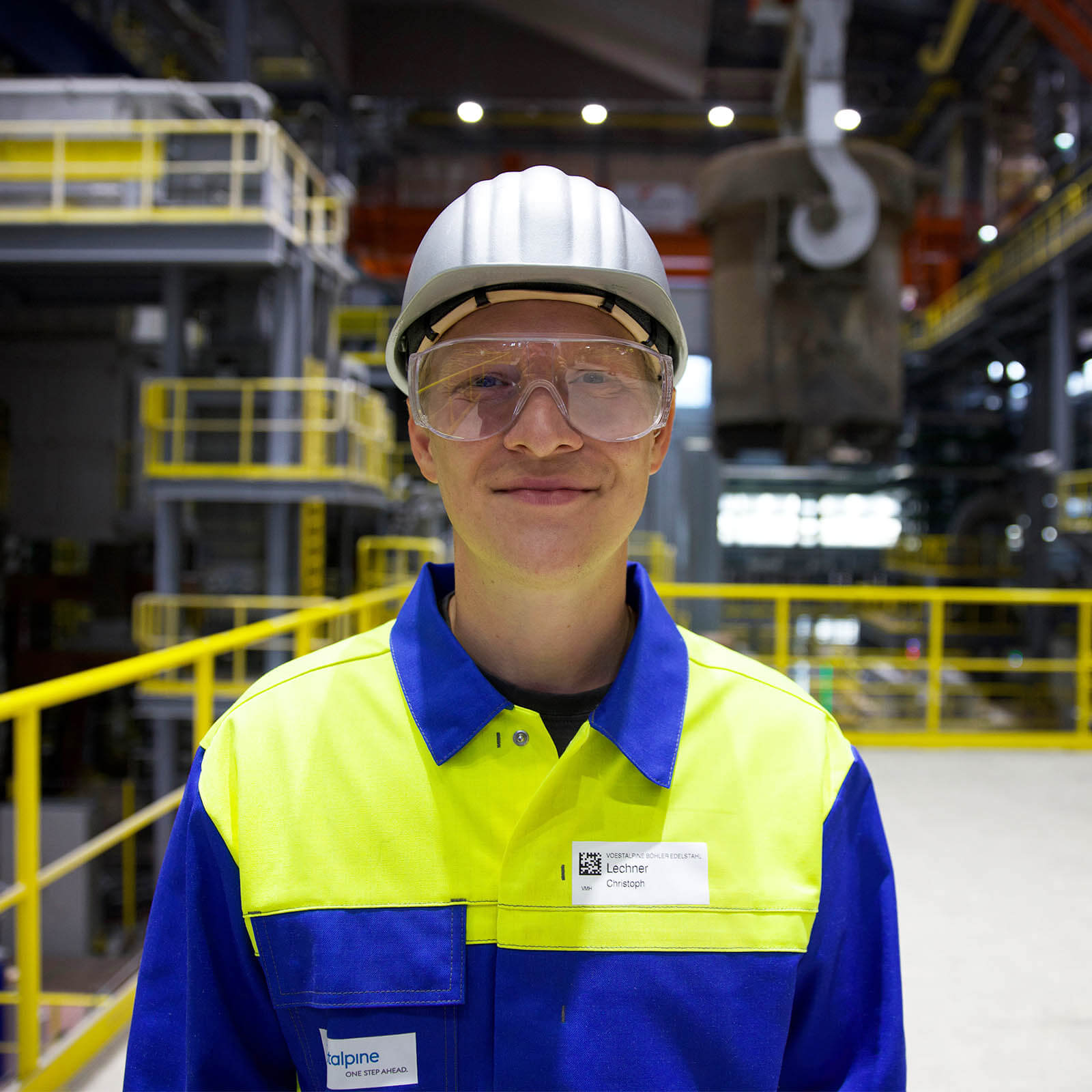 Christoph trägt blau-gelbe Arbeitskleidung, eine Schutzbrille und einen weißen Arbeitshelm und lächelt auf einer Plattform in einer Werkshalle in die Kamera
