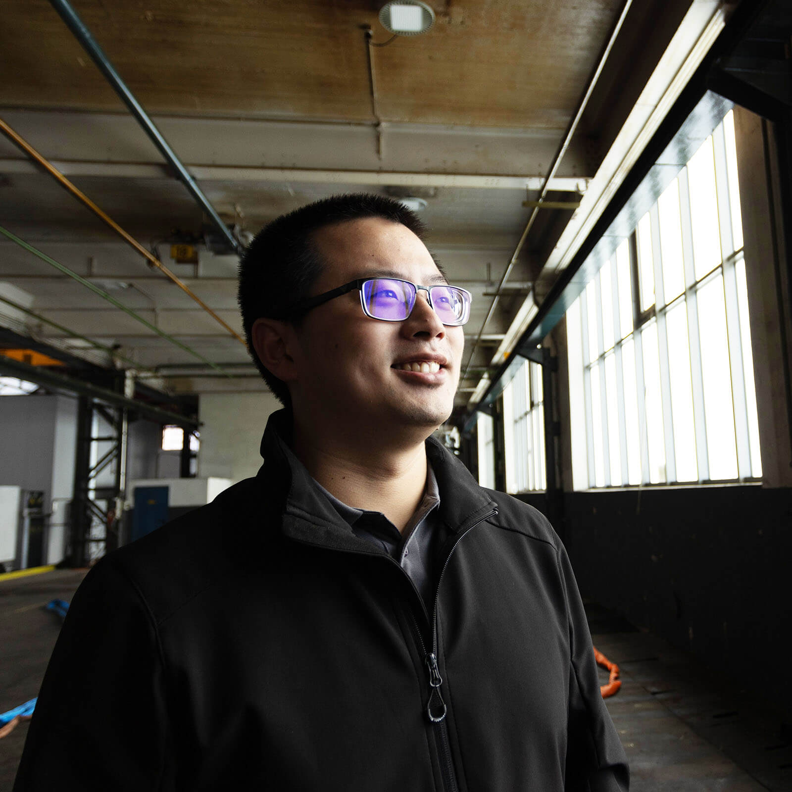 Liang, Mitarbeiter der voestalpine, lächelt in einer Werkshalle rechts richtung Fenster.