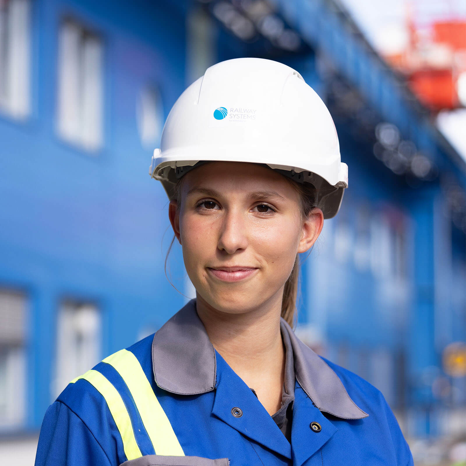 voestalpine Mitarbeiterin Lisa blickt mit einen weißen Schutzhelm und blaue Arbeitsbekleidung in die Kamera, im Hintergrund kann man Industriegebiet erkennen.