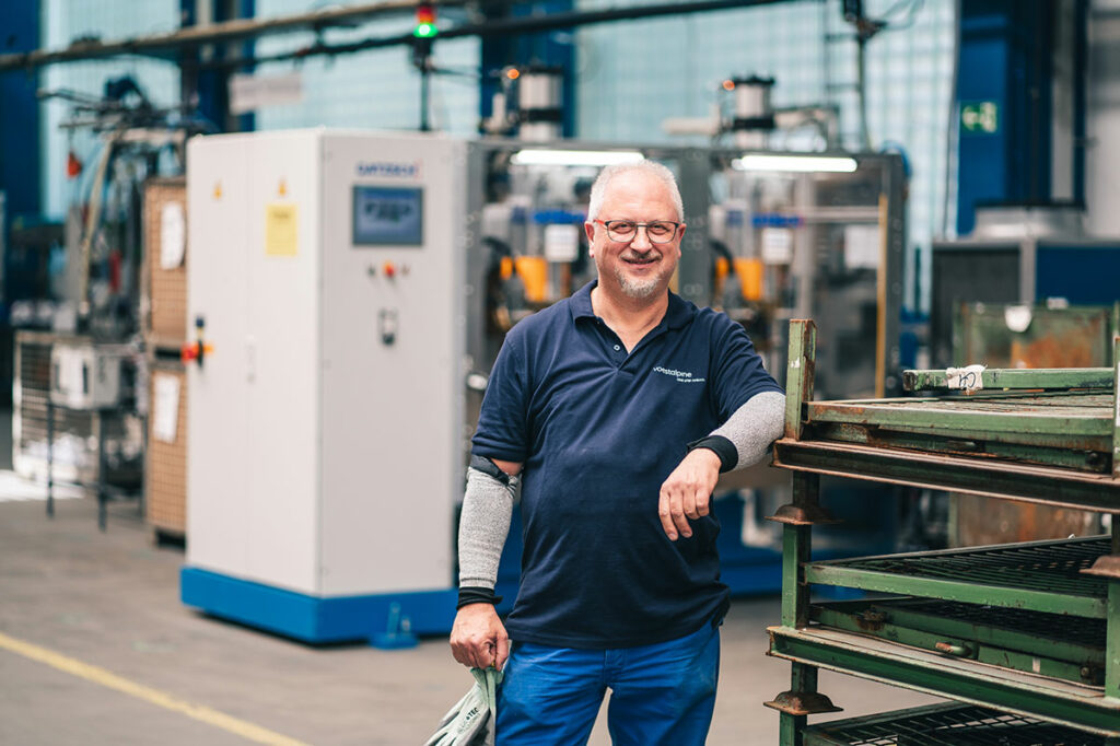 Bernd lächelt im Portrait in der Werkshalle an eine Metall Ablage gelehnt