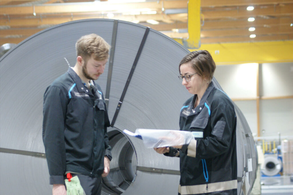 Angelika G. looks at a folder with an employee; large steel rolls lie in the background