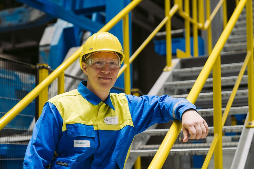 voestalpine employee Christoph leans against the yellow banister in his protective clothing in a production hall at voestalpine