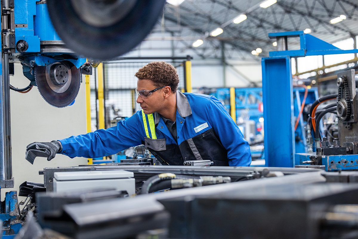 Nathan in blauer Schutzkleidung und Schutzbrille bedient eine große Industriemaschine in einer Produktionshalle. In der Hand hält er en Stück eines Stahlprofils.