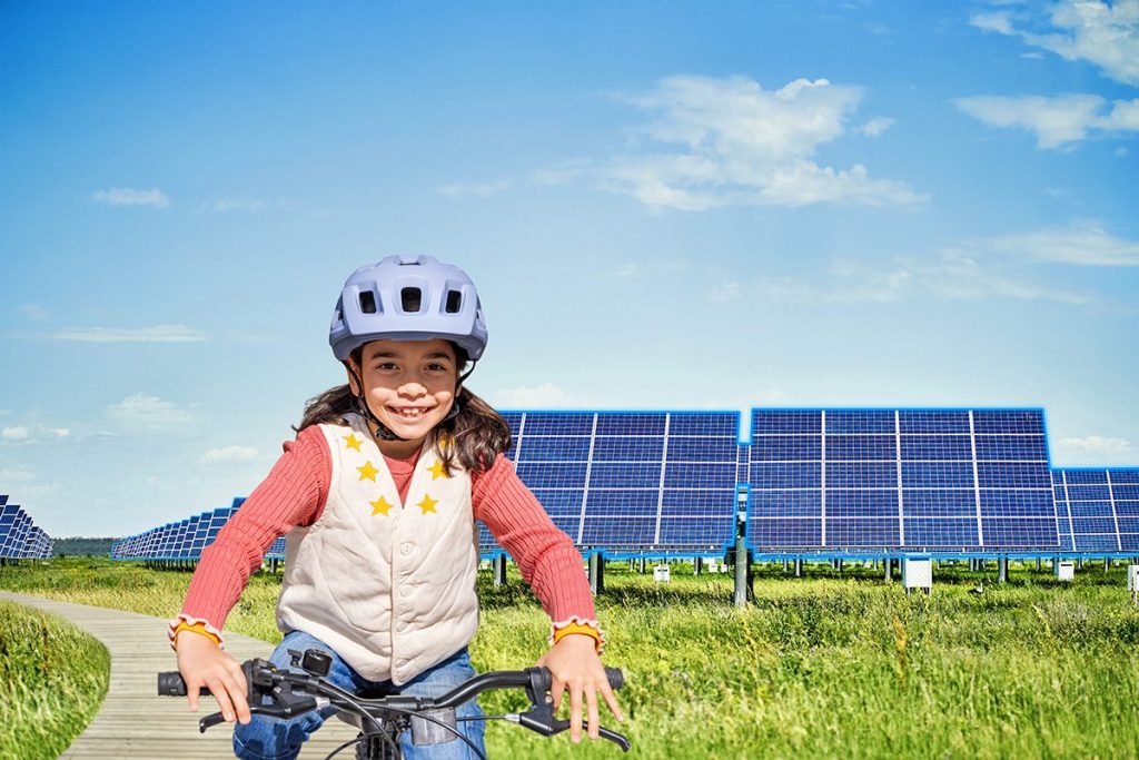 Ein lächelndes Kind mit Fahrradhelm fährt Fahrrad auf einem Holzweg, im Hintergrund sind große Solarpaneele auf einer grünen Wiese unter blauem Himmel zu sehen.