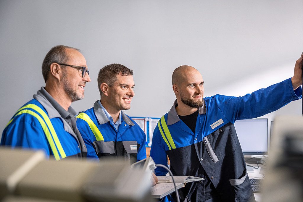 Drei voestalpine-Mitarbeiter in blauer Arbeitskleidung schauen gemeinsam auf einen Bildschirm. Einer zeigt mit dem Finger nach vorne, die Stimmung ist konzentriert und positiv.