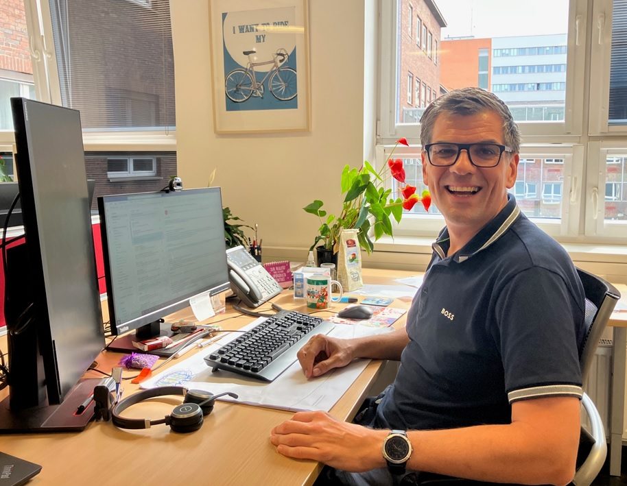 voestalpine Mitarbeiter Peter mit Brille und dunkelblauem Poloshirt sitzt lächelnd an einem Schreibtisch im Büro vor zwei Monitoren, mit Pflanzen und Fenstern im Hintergrund.