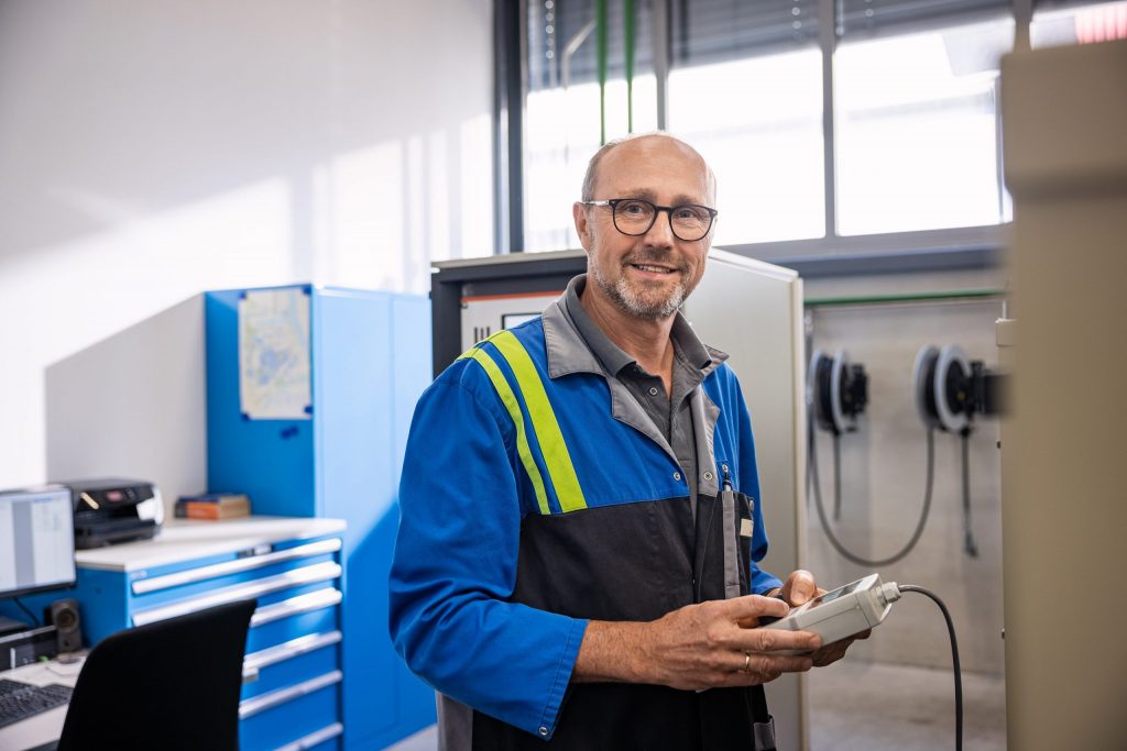 Portrait Kurt S., voestalpine-Forschungsleiter, er hält elektronisches Gerät in den Händen und lächest in die Kamera, in der Unschärfe im Hintergrund kann man sein Büro erkennen.