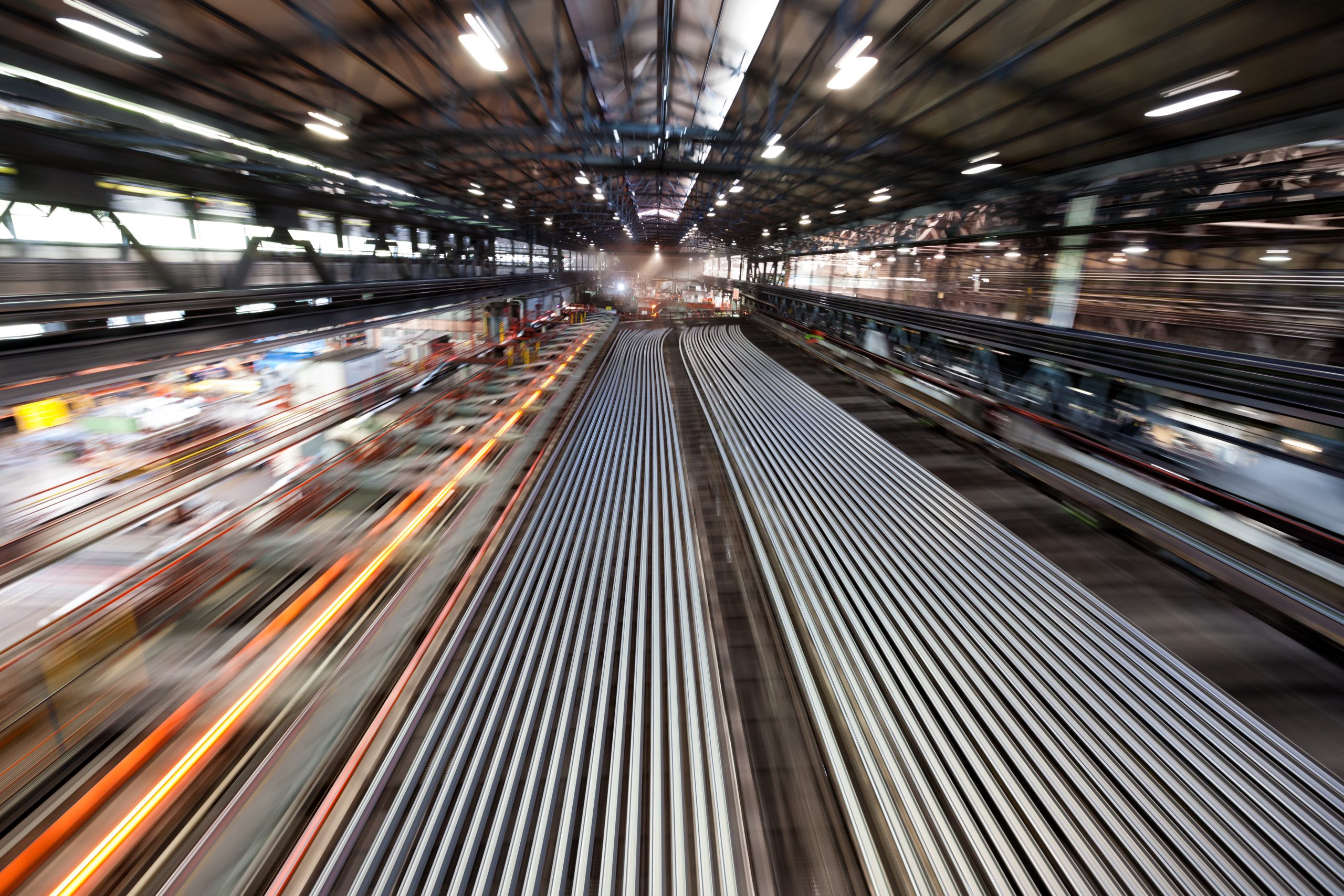 Blick in eine große Industriehalle einer voestalpine-Fertigungsanlage mit zahlreichen parallel liegenden, langen Schienenstahl-Profilen, die über den Boden zur Produktions- oder Lagerzone verlaufen; im Hintergrund technische Hallenstrukturen und Beleuchtung sichtbar.