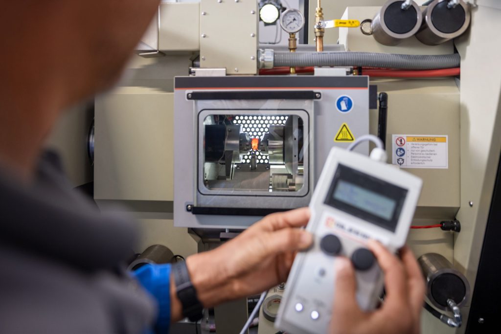 voestalpine Mitarbeiter elektronisches Gerät in den Händen und lächest in die Kamera, in der Unschärfe im Hintergrund kann man sein Büro erkennen.