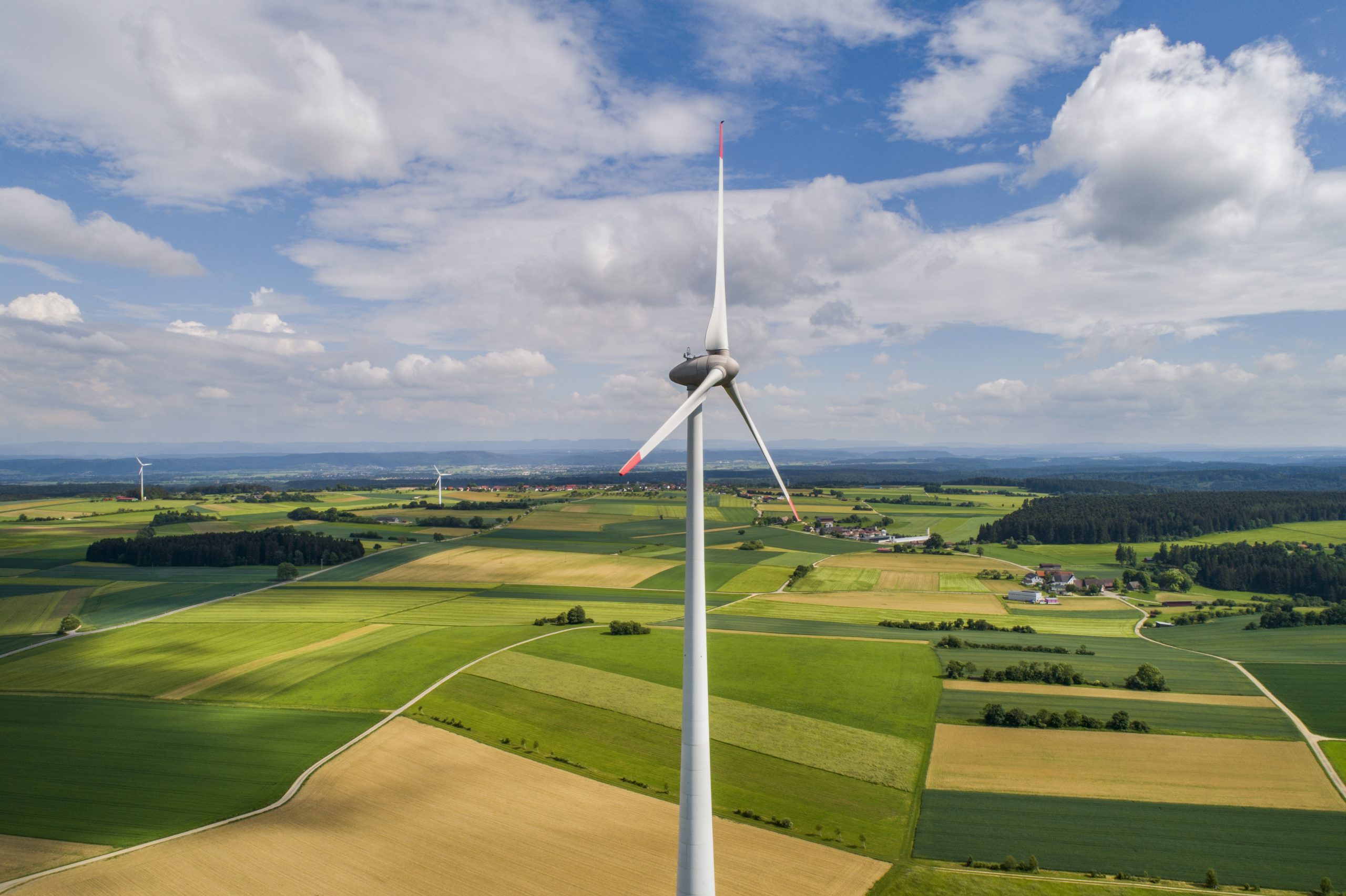 Große Windkraftanlage ragt über eine grüne, patchworkartige Agrarlandschaft mit Feldern, kleinen Dörfern und weiteren Windrädern unter einem teils bewölkten Himmel.