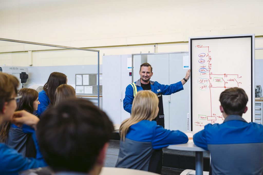 Instructor Christoph explains a technical circuit diagram on a whiteboard to a group of apprentices wearing blue work uniforms at a training center.