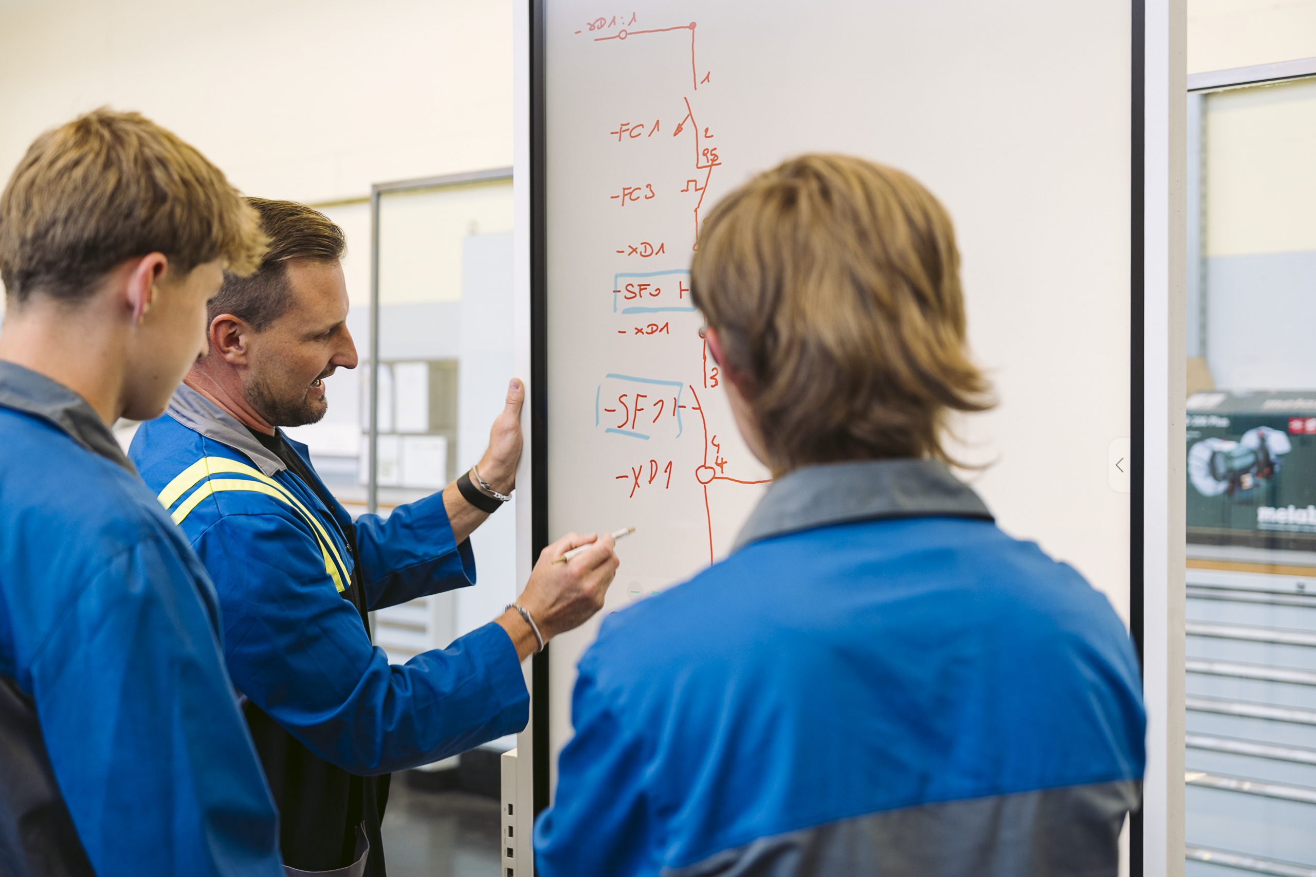 Instructor Christoph explains a technical circuit diagram to two apprentices on a whiteboard, adding details with a marker at a training center.