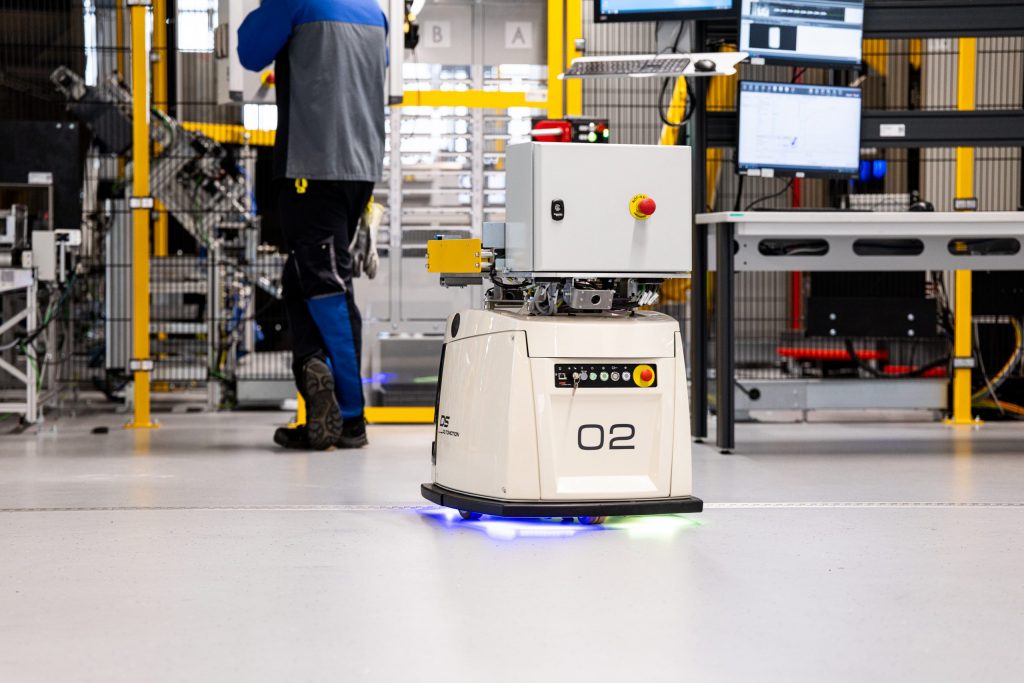 Autonomer mobiler Roboter (AMR) mit blauer LED-Beleuchtung fährt durch die moderne voestalpine Produktionshalle, im Hintergrund ein Mitarbeitender der voestalpine an einer Maschine.