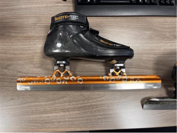 Nahaufnahme eines schwarzen Eisschnelllaufschuhs der Marke SKATE-TEC mit orange-silberner Kufe auf einem Holztisch.
