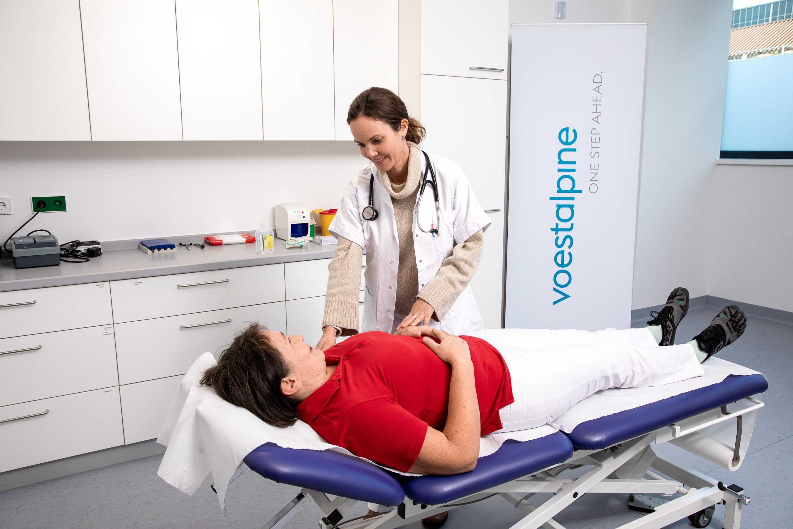 Pippa P. untersucht eine Patientin, die in einem Behandlungsraum auf einer Untersuchungsliege liegt; im Hintergrund stehen medizinische Geräte und ein Banner mit der Aufschrift „voestalpine – One Step Ahead“.