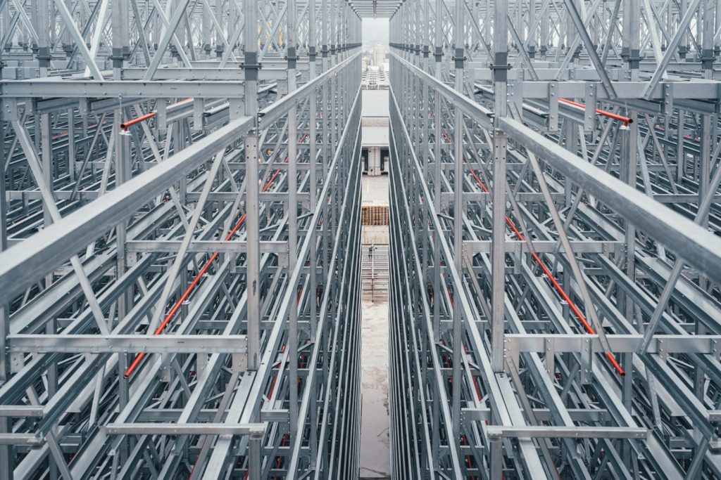 Innenansicht einer im Bau befindlichen Hochregalanlage aus Stahl mit symmetrischen Trägerstrukturen und schmalem Mittelgang.