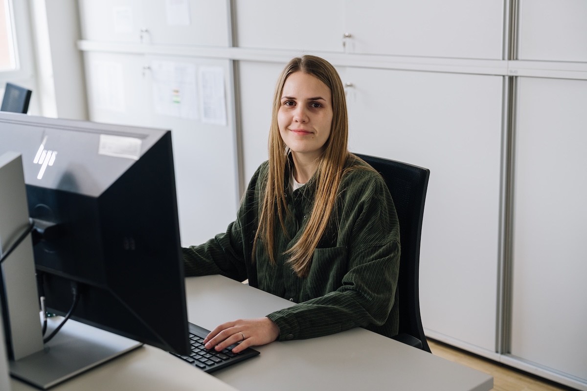 Patricia von voestalpine Krems sitzt an ihrem Arbeitsplatz vor zwei Monitoren und blickt lächelnd in die Kamera. Im Hintergrund sind helle Büroschränke zu sehen.