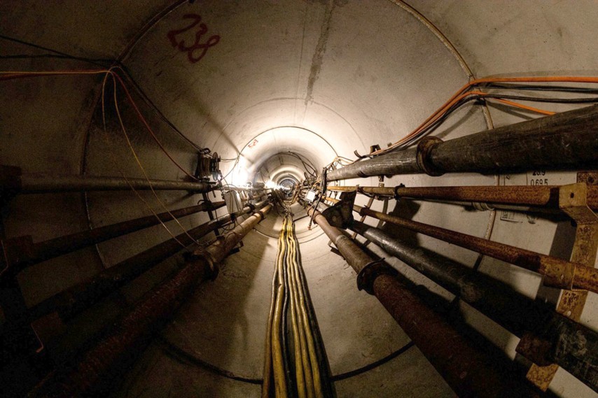 Blick durch einen langen, zylindrischen Versorgungstunnel aus Beton. Entlang der Tunnelwände verlaufen zahlreiche Rohre und Kabel, die perspektivisch auf einen hell erleuchteten Punkt am Ende des Tunnels zulaufen. Mehrere dicke Leitungen und rostige Rohrsysteme prägen das technische Erscheinungsbild. Die Aufnahme ist mittig ausgerichtet und erzeugt durch die starke Tiefenwirkung einen Eindruck von Länge und Enge.