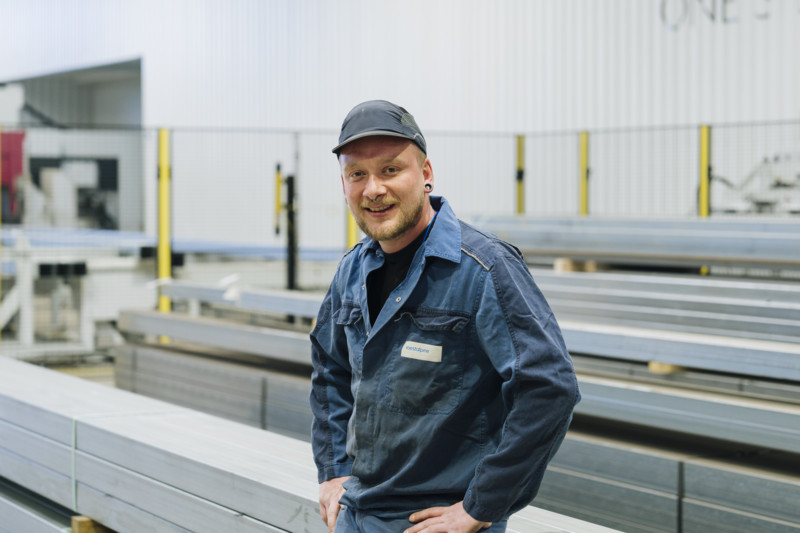 Wolfgang in Arbeitskleidung steht lächelnd zwischen gelagerten Stahlprofilen in einer Produktionshalle.