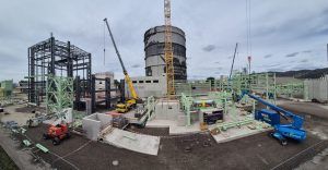 Weitläufige Industriebaustelle mit Stahlkonstruktionen, Kränen, Hubarbeitsbühnen und einem großen zylindrischen Industriegebäude im Hintergrund.