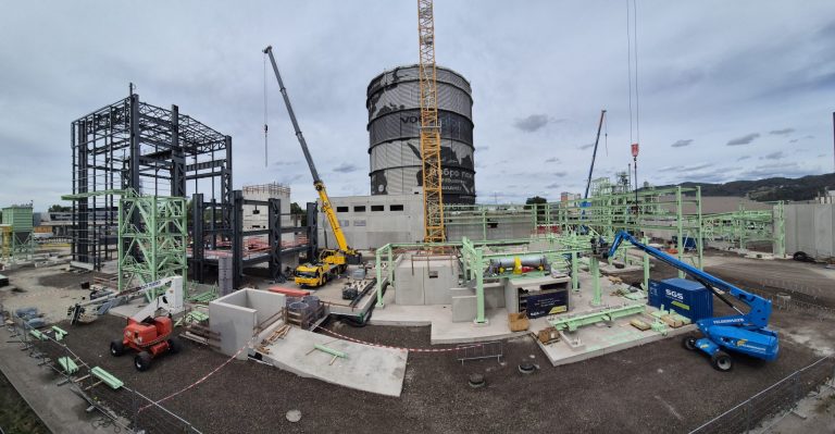 Weitläufige Industriebaustelle mit Stahlkonstruktionen, Kränen, Hubarbeitsbühnen und einem großen zylindrischen Industriegebäude im Hintergrund.