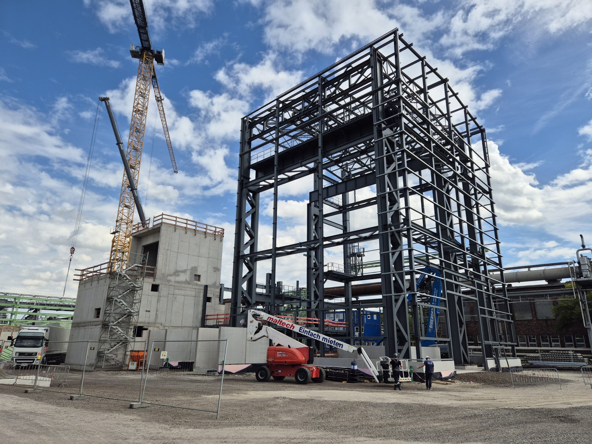 Mehrstöckige Stahlkonstruktion auf einer Industriebaustelle mit Betongebäude, Kränen und Hubarbeitsbühnen während der Montage.
