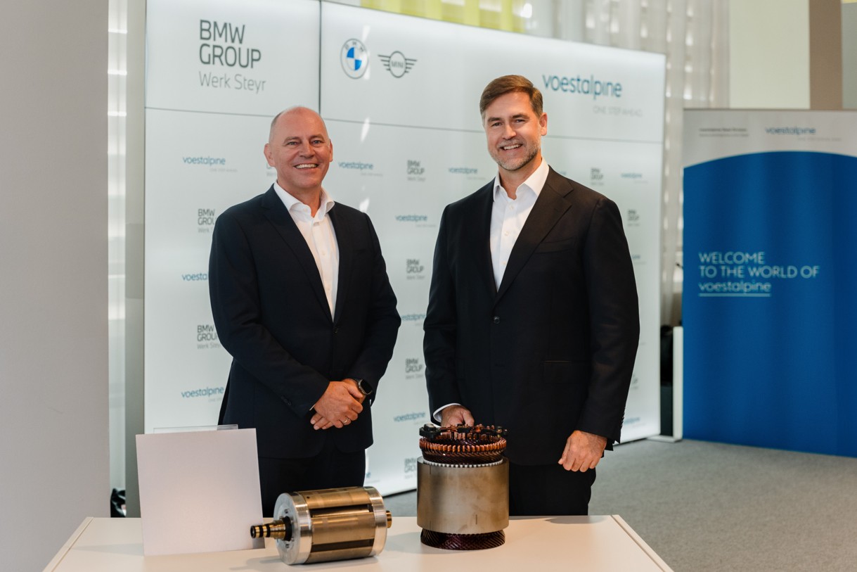 Zwei Unternehmensvertreter vor einer BMW Group und voestalpine Fotowand mit Komponenten eines Elektromotors auf einem Präsentationstisch.