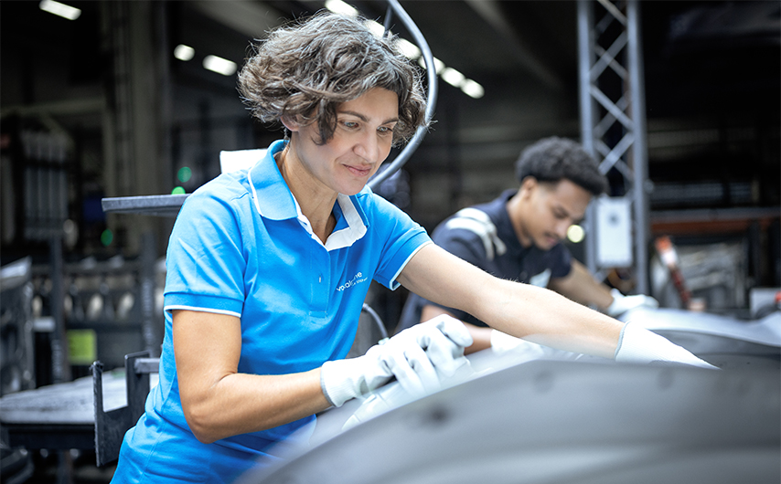 Employee in workwear and protective gloves working on a component in a production hall, with a colleague working in the background.
