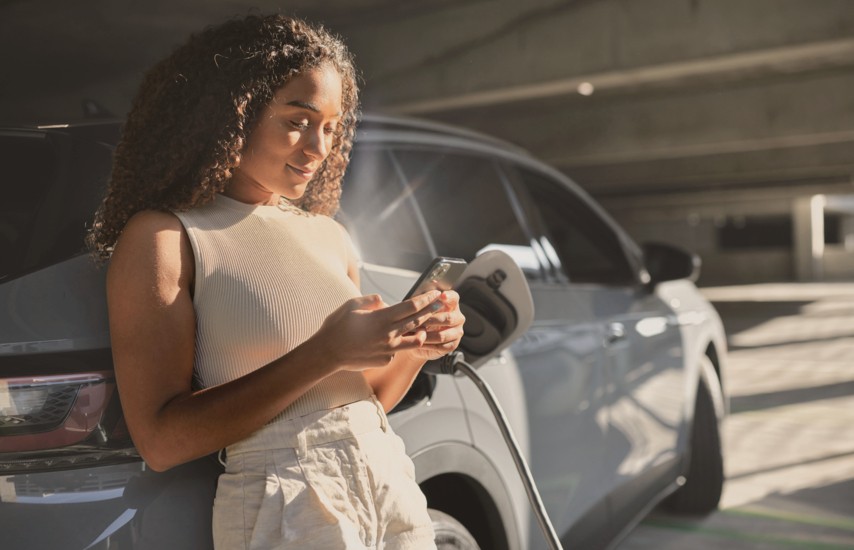 Frau steht mit Smartphone neben einem Elektroauto und lädt das Fahrzeug an einer Ladestation in einem modernen Parkhaus.