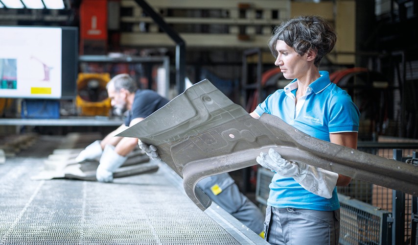 Employee in workwear and protective gloves inspecting a large automotive body component in a production hall.