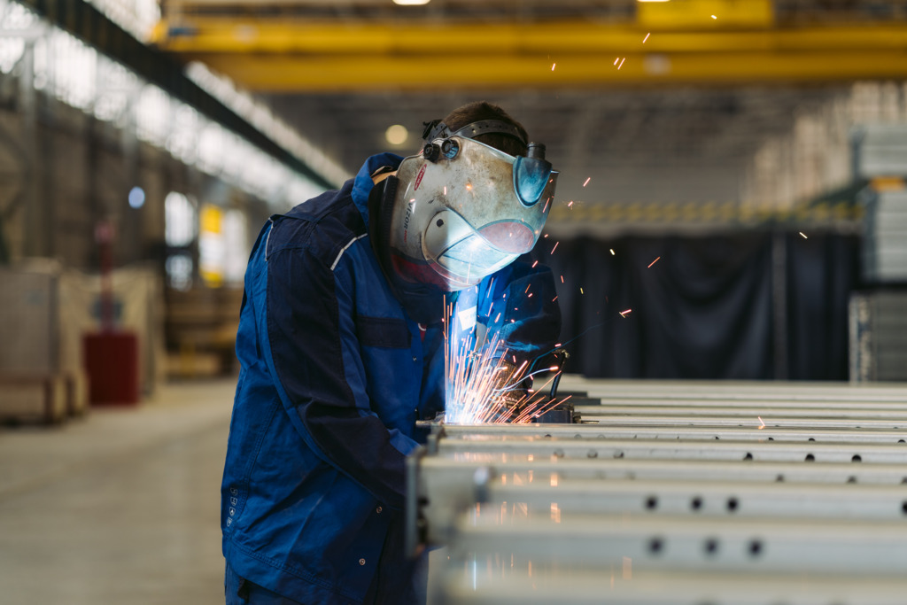 Manuel in blauer Arbeitskleidung und mit Schutzhelm schweißt Metallprofile in einer Produktionshalle. Dabei entstehen helle Lichtbögen und zahlreiche Funken.