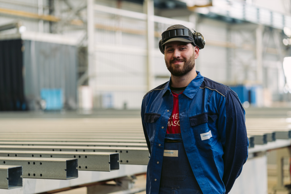 Manuel, Schweiss-Techniker bei der voestalpine, steht in blauer Arbeitskleidung und mit Schutzkappe samt Gehörschutz steht in einer Produktionshalle neben mehreren langen Metallprofilen.