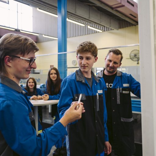 Instructor Christoph supports two apprentices during a practical exercise at the training center while one student presents a workpiece and others watch attentively.