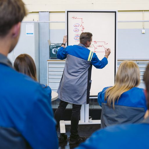 Instructor Christoph explains a technical circuit diagram to a group of apprentices on a whiteboard, adding details to the schematic.