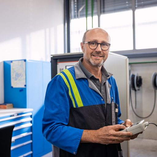 Portrait of Kurt S., head of research at voestalpine, holding an electronic device in his hands and smiling at the camera, with his office visible in the blurred background.