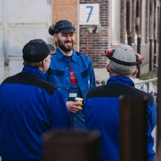 Manuel und zwei Kollegen von der voestalpine blauer Arbeitskleidung und Schutzkappen unterhalten sich während einer Pause vor einem Backsteingebäude. Manuel in der Mitte hält einen Kaffeebecher in der Hand.
