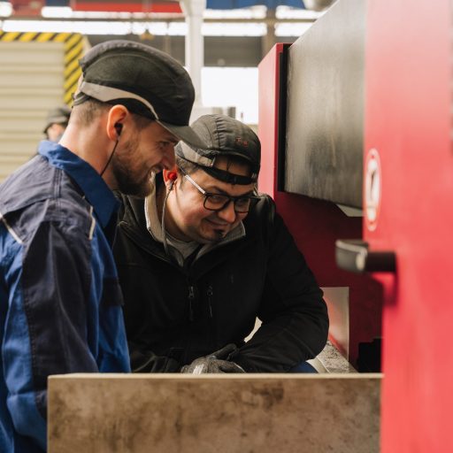 Daniel und ein voestalpine Kollege mit Schutzbrillen und Arbeitskleidung besprechen einen Arbeitsschritt an einer Industriemaschine in einer modernen Fertigungsumgebung.