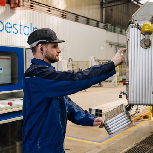 Mitarbeiter mit Schutzhelm und Arbeitskleidung steuert eine Fertigungsanlage über ein Bedienfeld in einer modernen Produktionsumgebung.