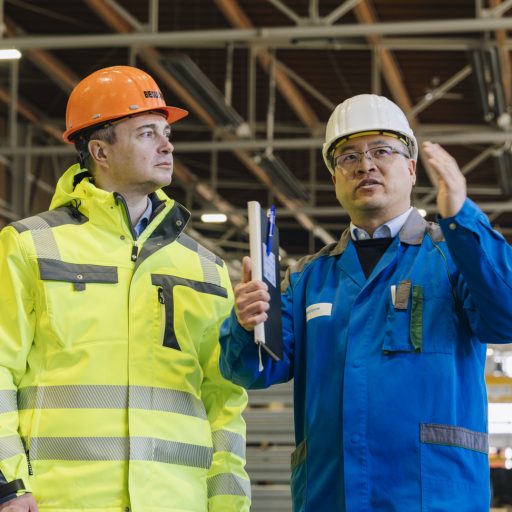 Zwei Mitarbeiter in Schutzkleidung tauschen sich während eines Rundgangs über technische Abläufe und Produktionsprozesse in einer Industriehalle aus.