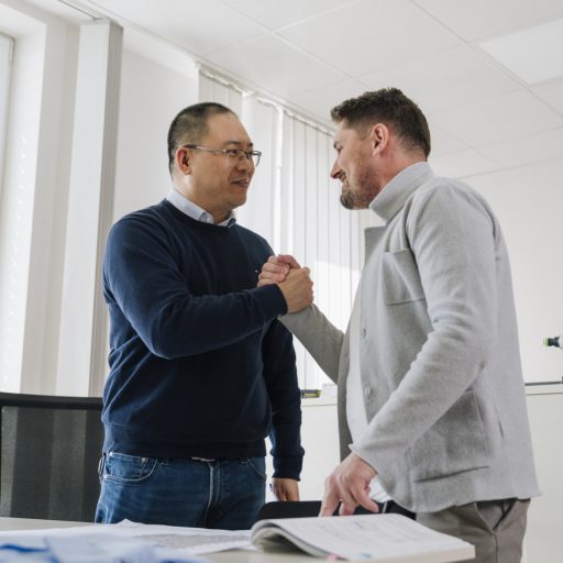 Hairuos und ein weiterer voestalpine Mitarbeiter stehen an einem Arbeitsplatz in einem modernen Büro, begrüßen sich per Handschlag und führen ein persönliches Gespräch.