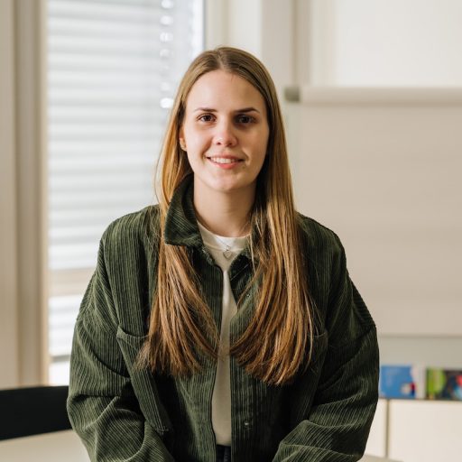 Patricia von voestalpine Krems sitzt in einem Besprechungsraum und blickt lächelnd in die Kamera. Im Hintergrund stehen ein Flipchart und ein Tisch.