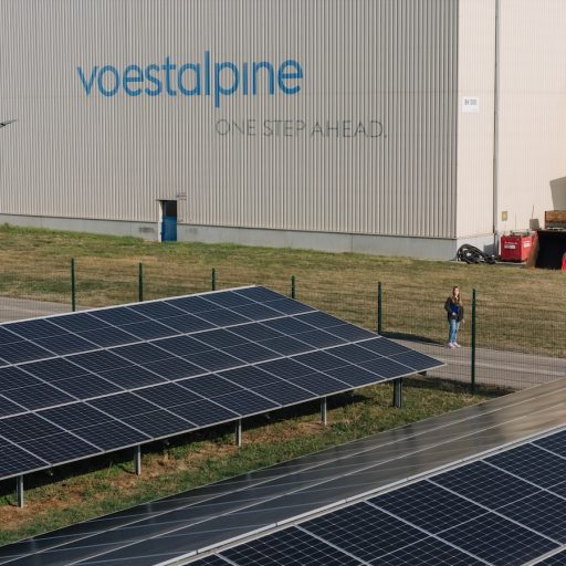 A solar power system in front of the production halls at voestalpine Krems under a blue sky. The words “voestalpine – one step ahead” are visible on one of the factory buildings. Just barely visible between the building and the solar power system is Patricia, who is looking toward the camera from a distance.