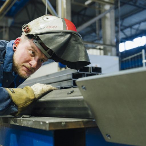 Wolfgang in workwear with his welding helmet raised inspects a steel component in a production hall.