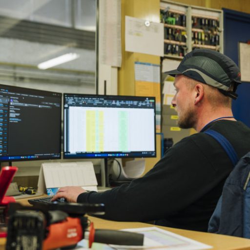 Wolfgang at an office workstation in the production area working on two monitors displaying digital overviews and tables.