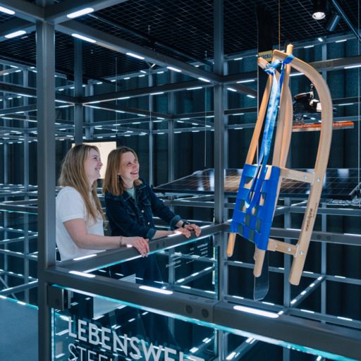 Zwei Besucherinnen stehen im begehbaren Grid der voestalpine Stahlwelt und sehen sich eine Rodel als Exponat an. Die Inszenierung zeigt, wie Stahl in unterschiedlichen Lebensbereichen und Alltagsprodukten zum Einsatz kommt.