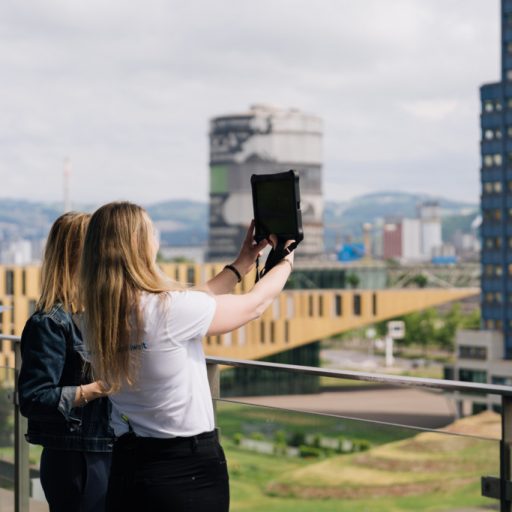 Zwei Besucherinnen auf der Terrasse der voestalpine Stahlwelt blicken mit einem hochgehaltenen Tablet auf das Werksgelände in Linz.