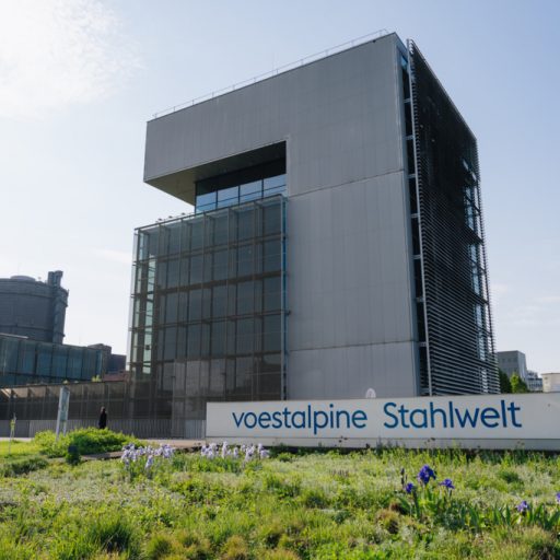 Außenansicht der voestalpine Stahlwelt in Linz mit moderner Glas- und Stahlfassade, begrüntem Vorplatz und sichtbarem Schriftzug am Gebäude.