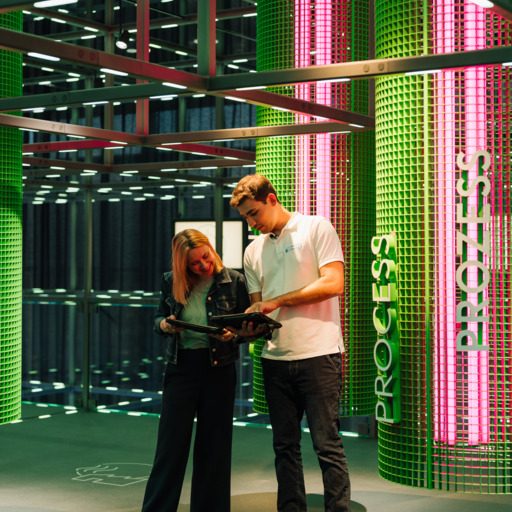 Zwei Besucher:innen stehen in der voestalpine Stahlwelt unter dem beleuchteten Grid und informieren sich mit einem Tablet über die Ausstellung. Im Hintergrund sind farbig inszenierte Bereiche zu Prozessen der Stahlproduktion zu sehen.