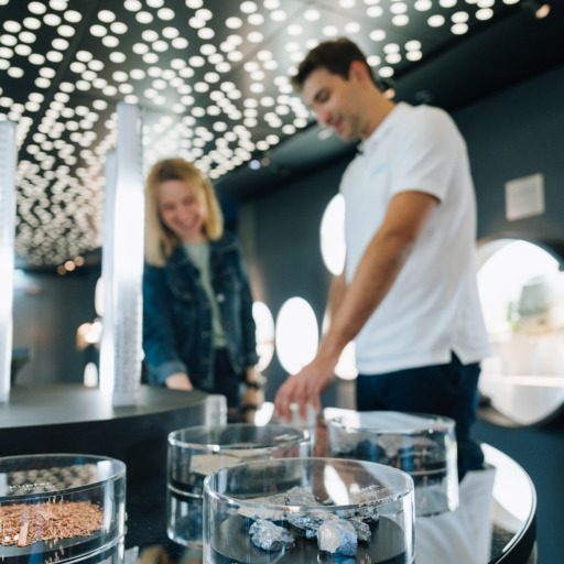 Zwei Besucher:innen stehen in der voestalpine Stahlwelt vor runden Glasvitrinen mit Rohstoffen und Materialien der Stahlproduktion. Die Lichtinszenierung an der Decke unterstreicht den modernen Ausstellungscharakter.